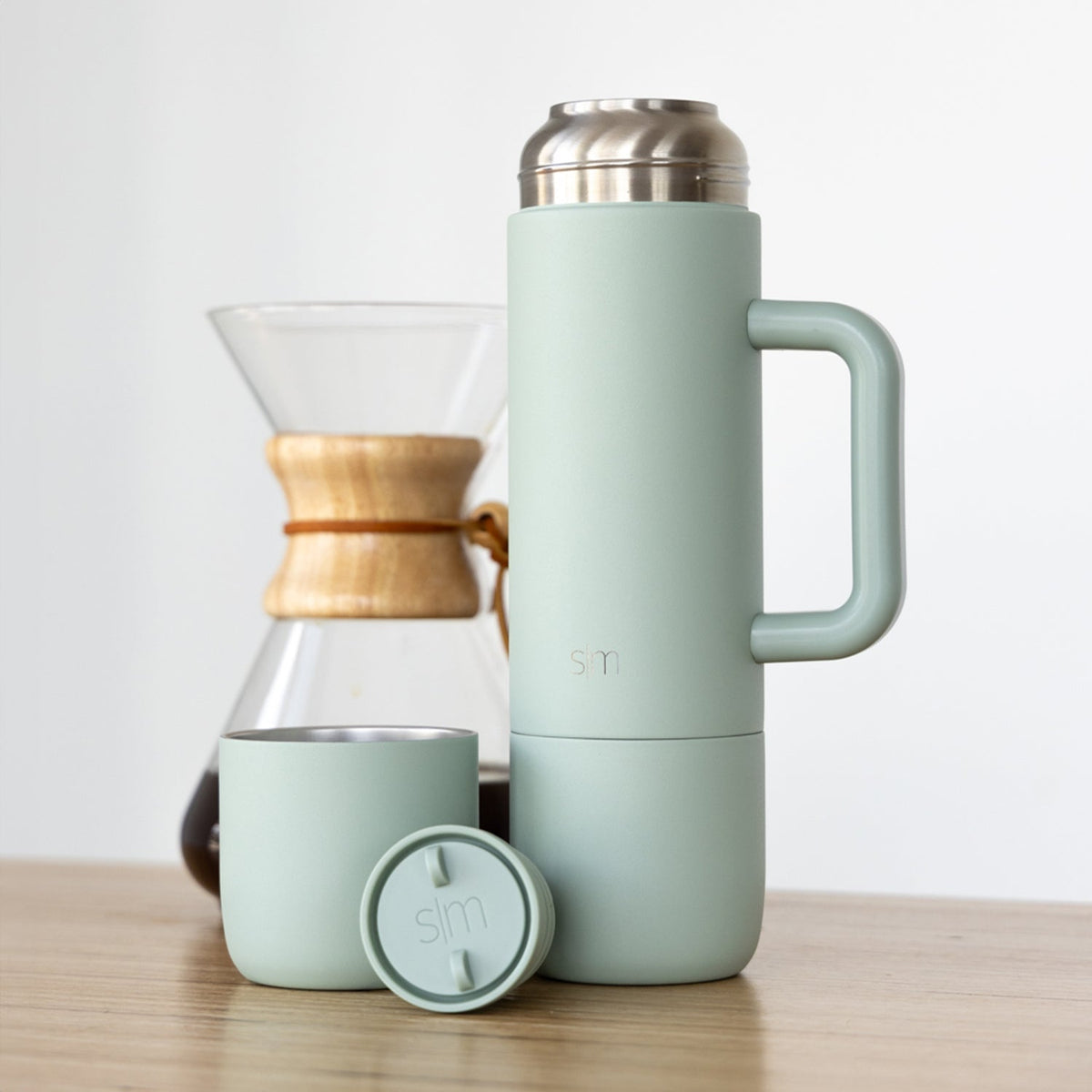 Sage green insulated tumbler and matching cup with warm wood and silver metallic accents on wooden surface